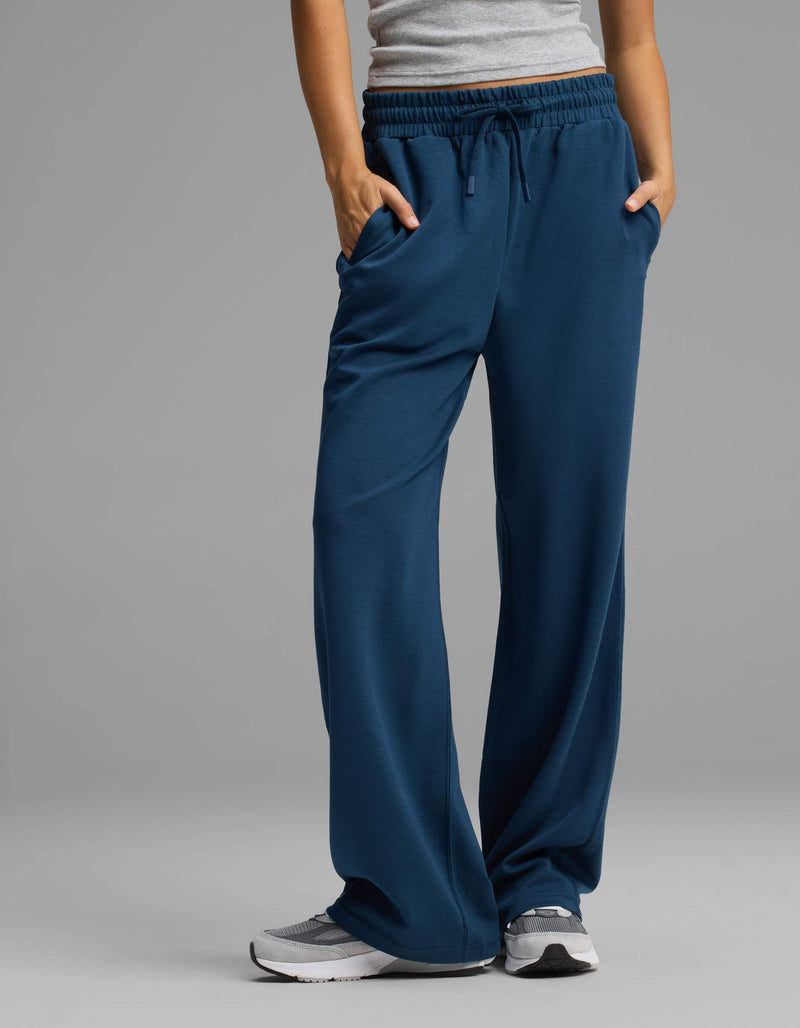 A person is wearing the Cozy Earth Women's Versa Straight Leg Pant in blue, paired with a gray tank top and gray sneakers, standing with hands in their pockets against a plain gray background.