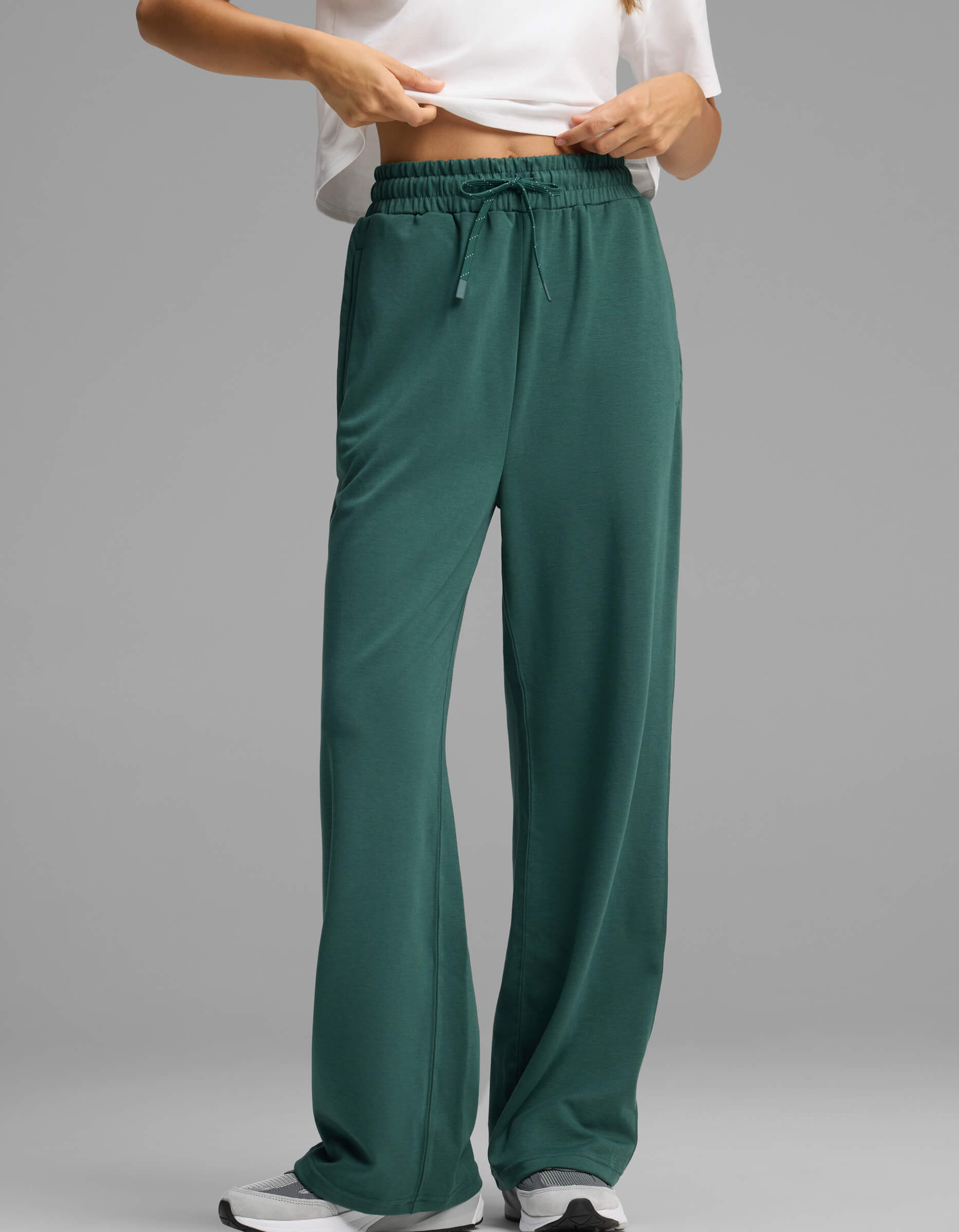 A model showcases the lower half of the Women's Versa Straight Leg Pant by Cozy Earth in green, paired with a white shirt and white sneakers, standing against a plain grey background and seen adjusting the waistband.