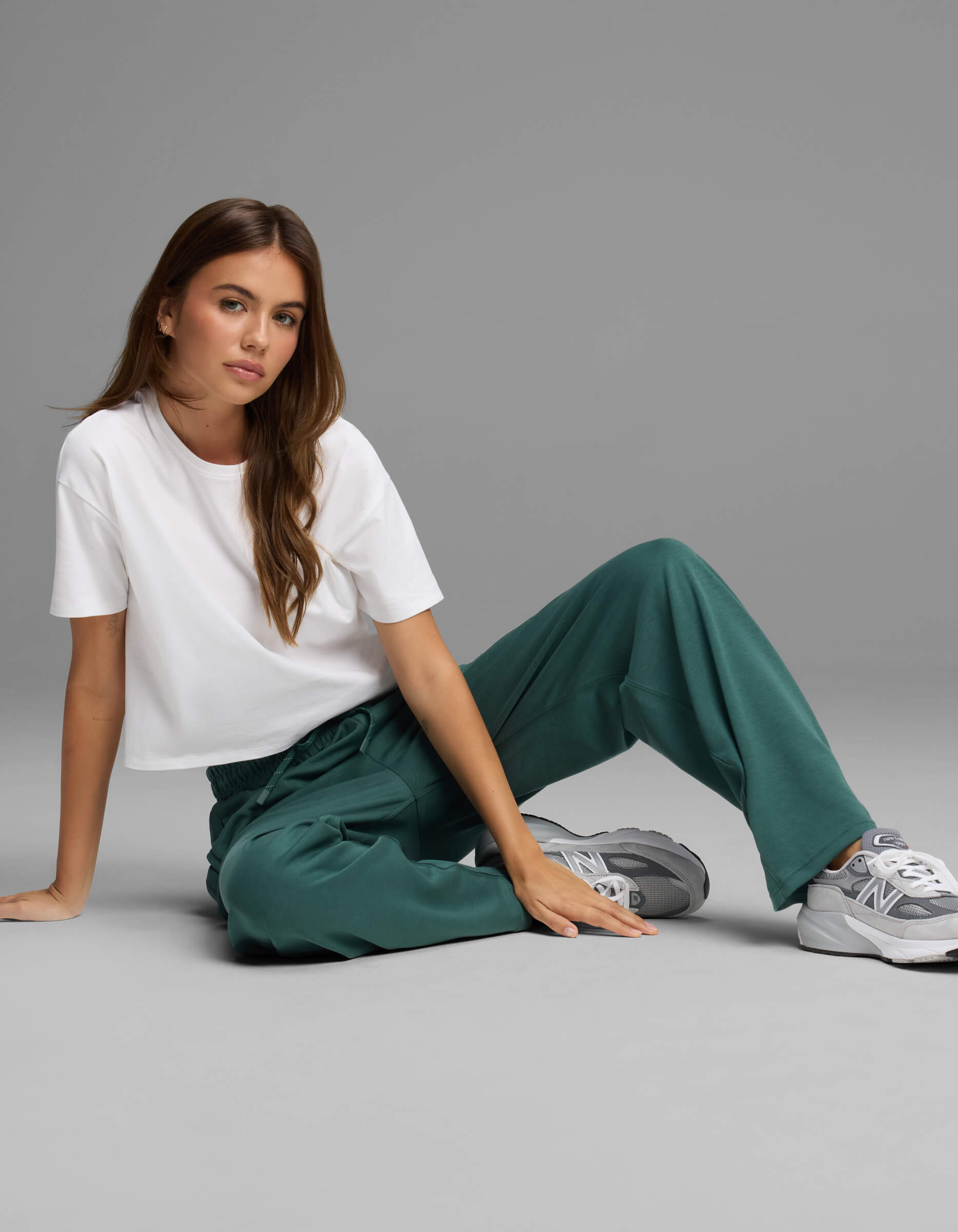A woman with long brown hair sits on the floor, wearing a white t-shirt, Cozy Earth Women's Versa Straight Leg Pant in green, and gray sneakers, posing against a plain gray background.