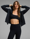 A woman poses against a plain gray background wearing the Cozy Earth Women's Versa Full-Zip Hoodie in black, styled with matching black sports bra and leggings, as she lifts the hood over her head.|Color:Jet Black