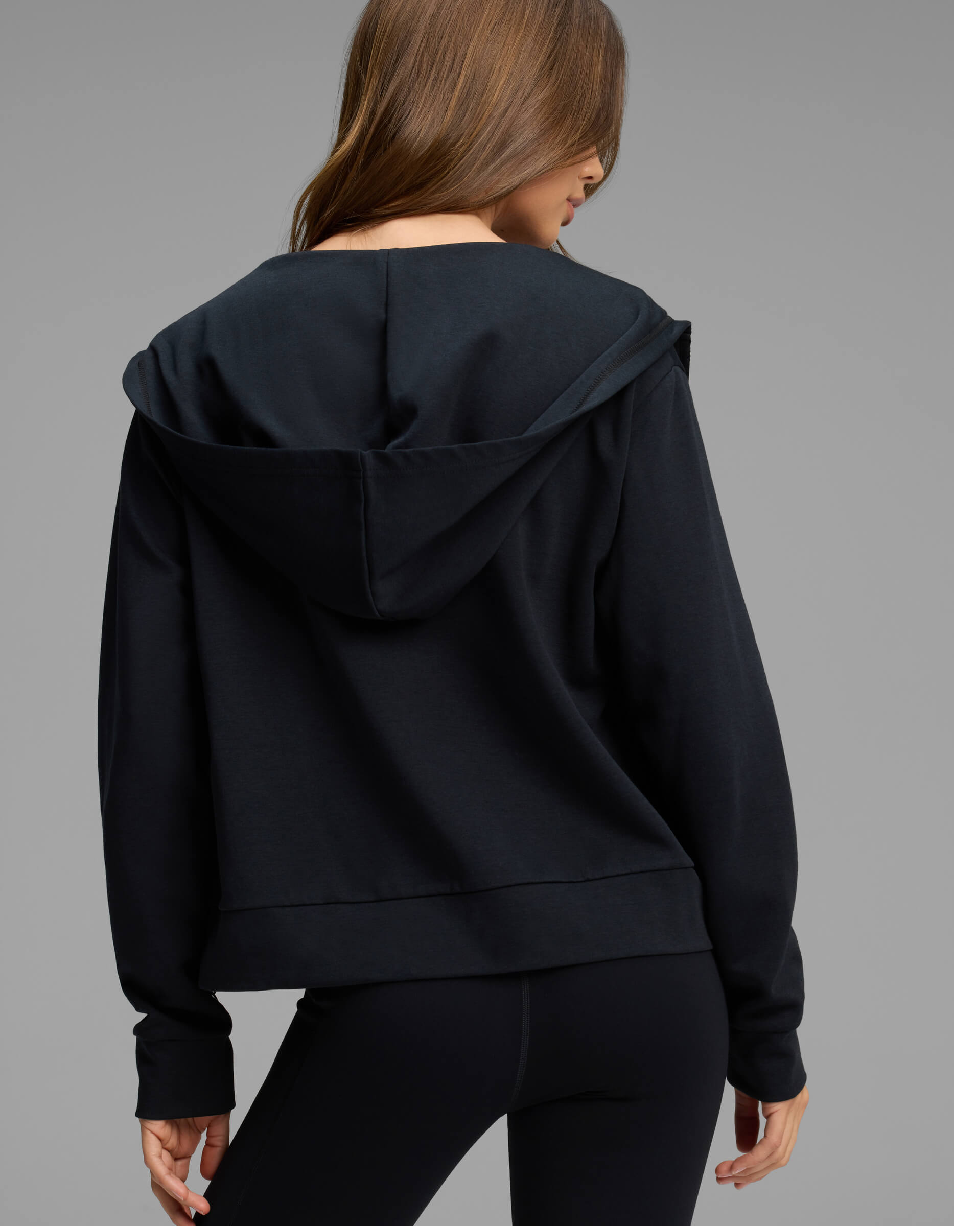 A person with long brown hair wears the Cozy Earth Women's Versa Full-Zip Hoodie in black, paired with black leggings, and stands facing away from the camera against a plain gray background.|Color:Jet Black