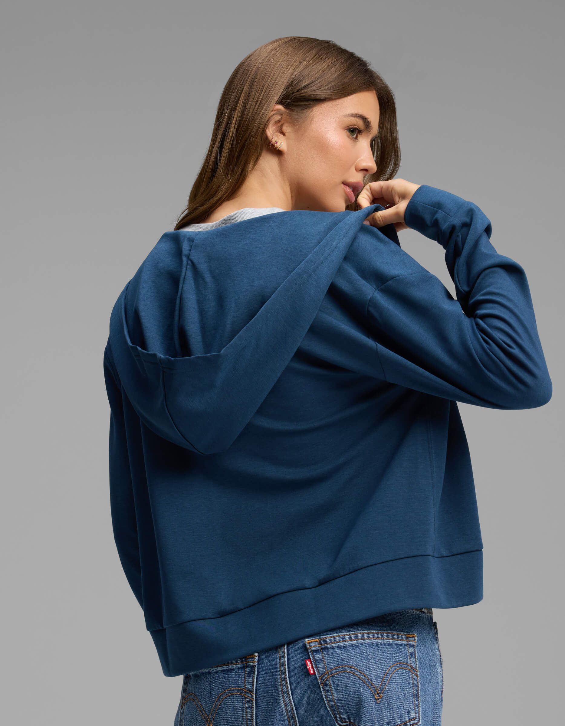 A woman with long brown hair wears the Cozy Earth Women's Versa Full-Zip Hoodie in blue and jeans, looking over her shoulder while holding the collar. She stands against a plain gray background.