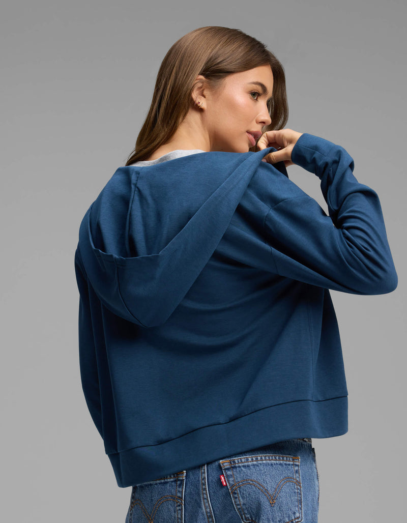 A woman with long brown hair wears the Cozy Earth Women's Versa Full-Zip Hoodie in blue and jeans, looking over her shoulder while holding the collar. She stands against a plain gray background.