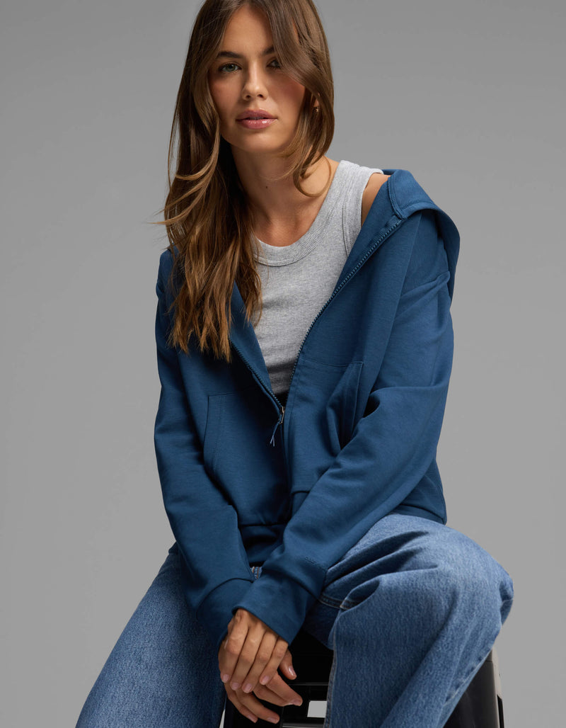 A woman with long brown hair wearing the Cozy Earth Women's Versa Full-Zip Hoodie in blue, along with a gray t-shirt and blue jeans, sits on a stool against a plain gray background, looking at the camera.