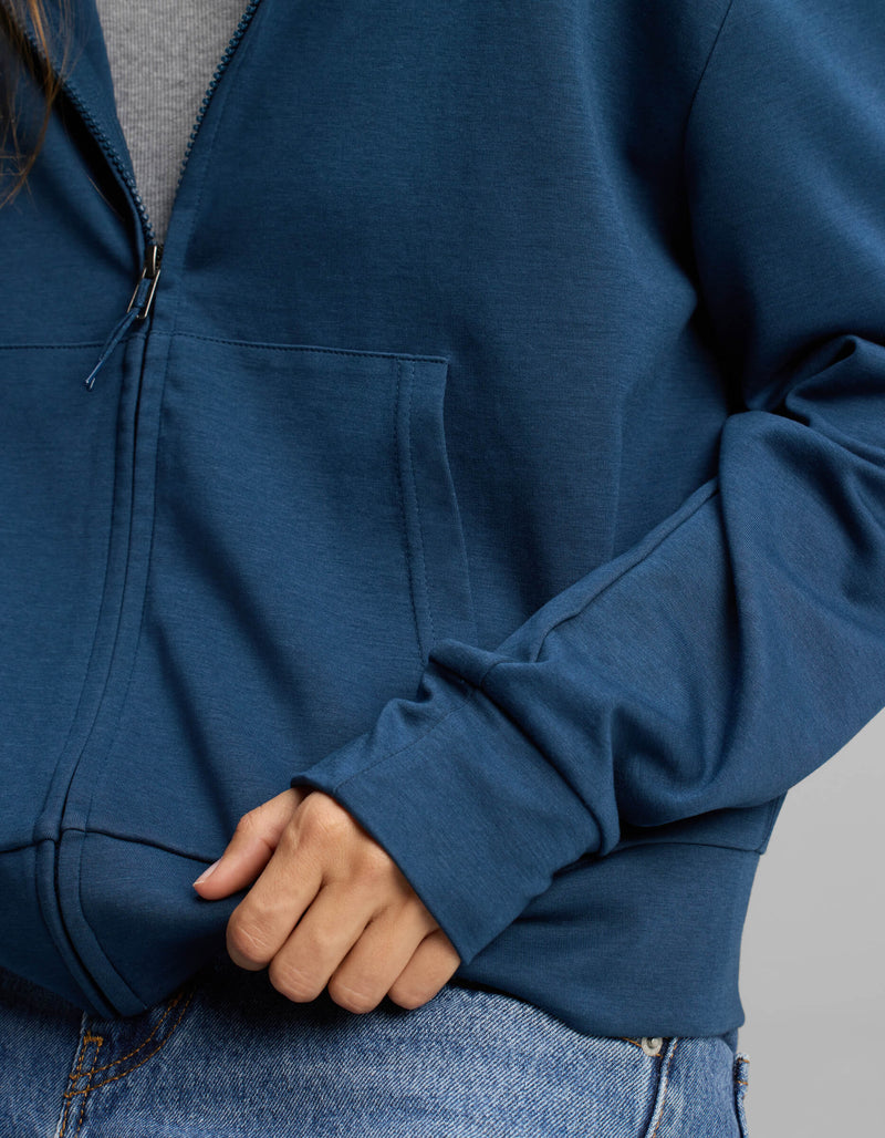 A person is shown from the chest down wearing Cozy Earth's Women's Versa Full-Zip Hoodie in blue and blue jeans, with one hand holding the hem of the hoodie and the other tucked into a pocket.