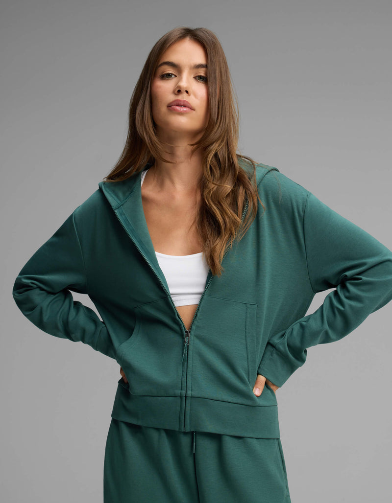 A woman with long brown hair stands with her hands on her hips against a plain gray background, wearing the Cozy Earth Women's Versa Full-Zip Hoodie in green over a white crop top.