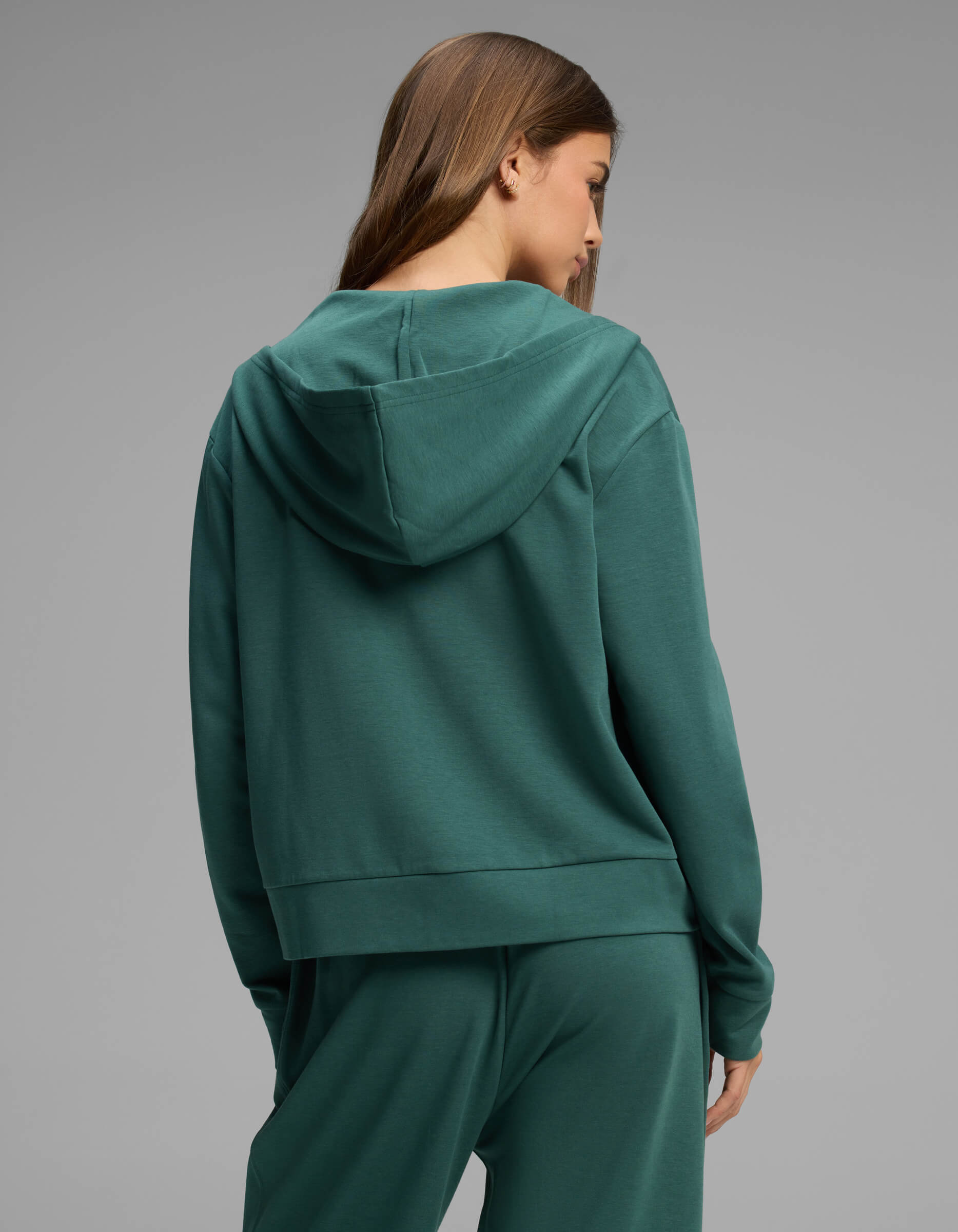 A woman with long brown hair is seen from behind against a plain gray background, wearing the Cozy Earth Women's Versa Full-Zip Hoodie in green with matching pants.