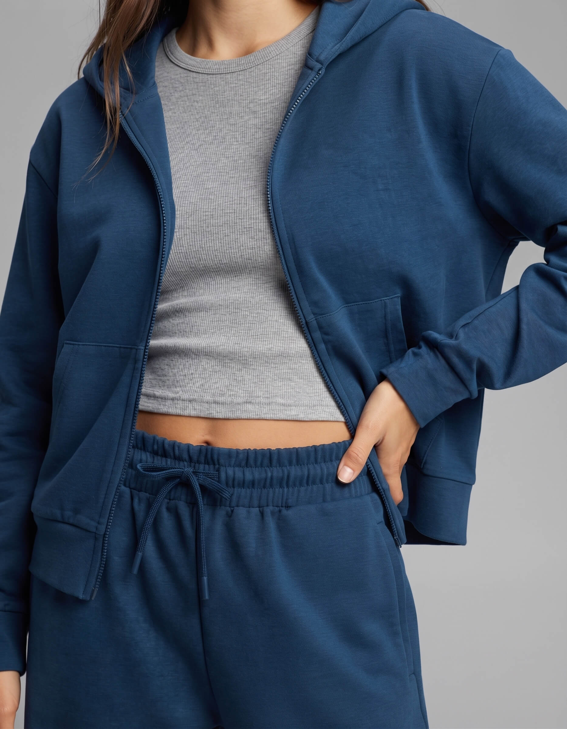 A person is wearing the Cozy Earth Women's Versa Zip-Down Hoodie & Straight Leg Pant Set in blue, paired with a light gray ribbed crop top. Their hand rests in the hoodie pocket, and the background is plain gray.