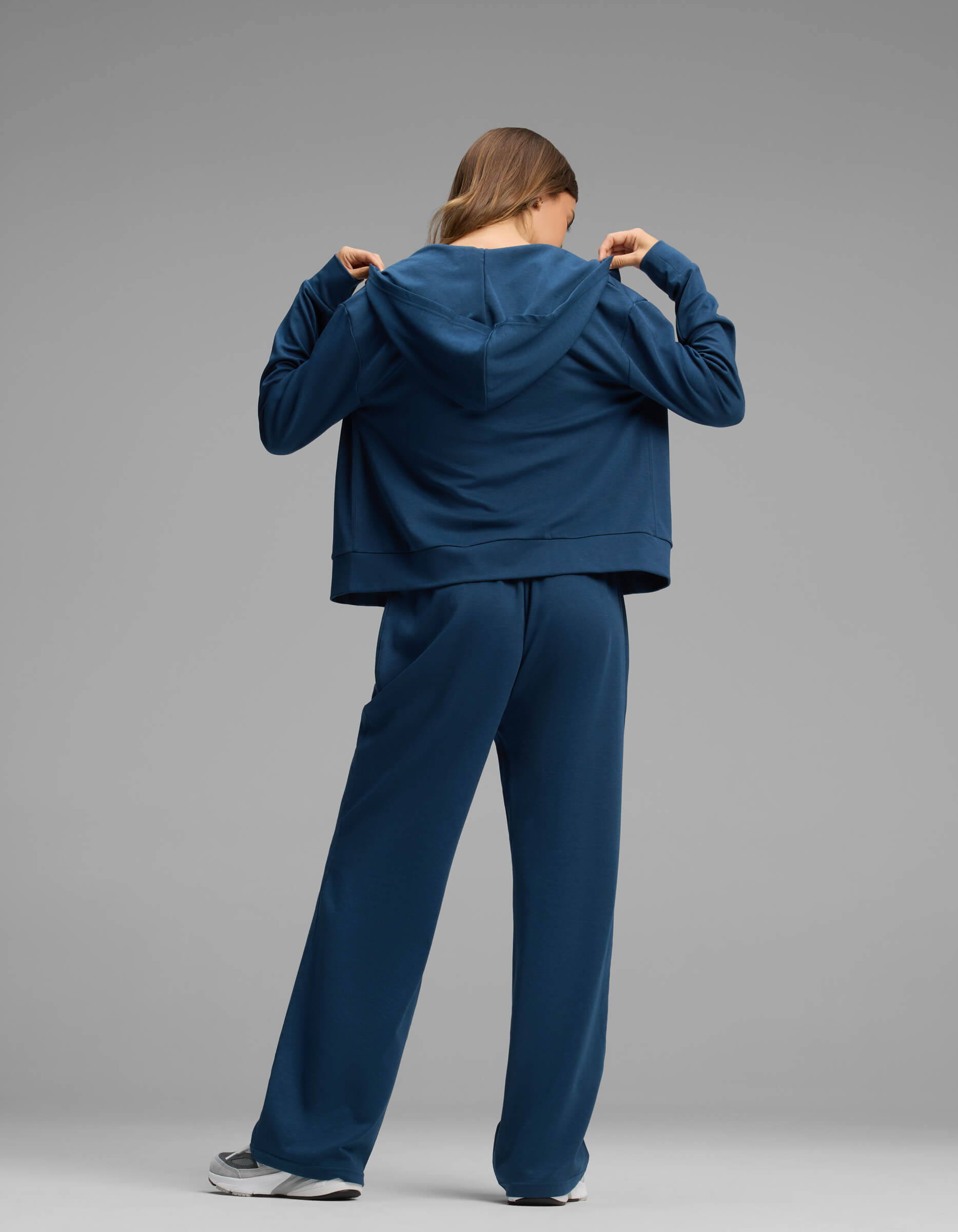 A woman stands facing away, wearing the Cozy Earth Women's Versa Zip-Down Hoodie & Straight Leg Pant Set in blue. She lifts the hoodie by the collar with both hands against a plain gray background.