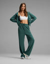 A woman poses in the Cozy Earth Women's Versa Zip-Down Hoodie & Straight Leg Pant Set, featuring a green hue, paired with a white sports bra and white sneakers, standing against a plain gray background.|Color:Uniform