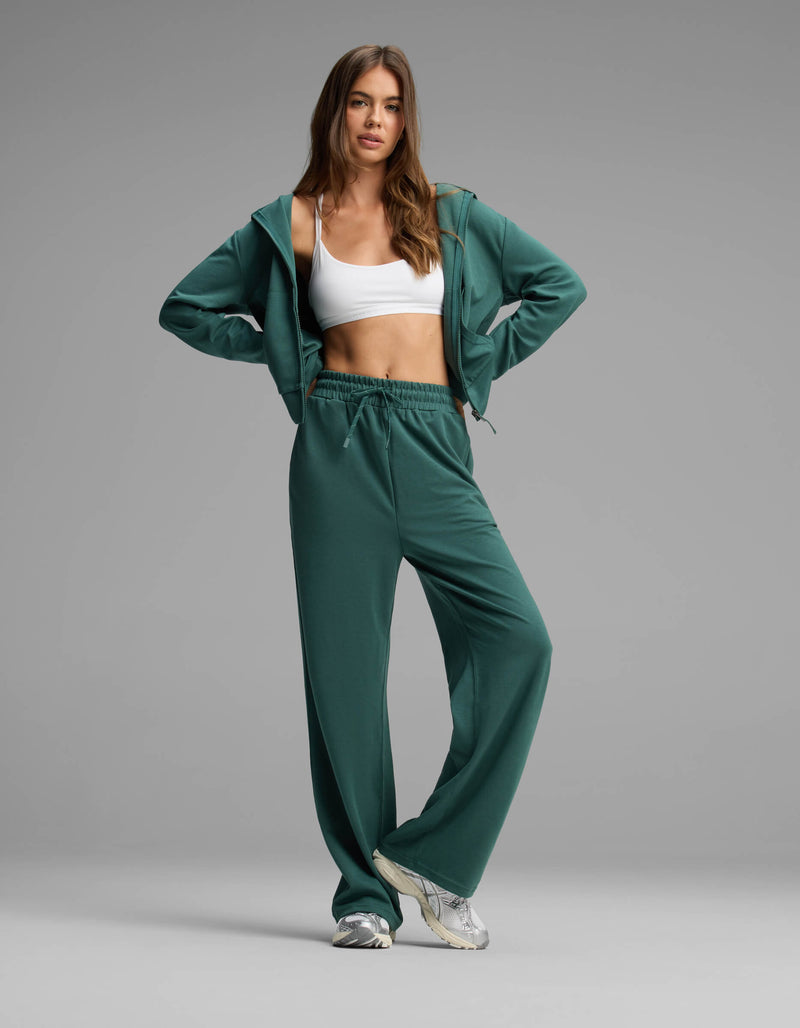 A woman poses in the Cozy Earth Women's Versa Zip-Down Hoodie & Straight Leg Pant Set, featuring a green hue, paired with a white sports bra and white sneakers, standing against a plain gray background.