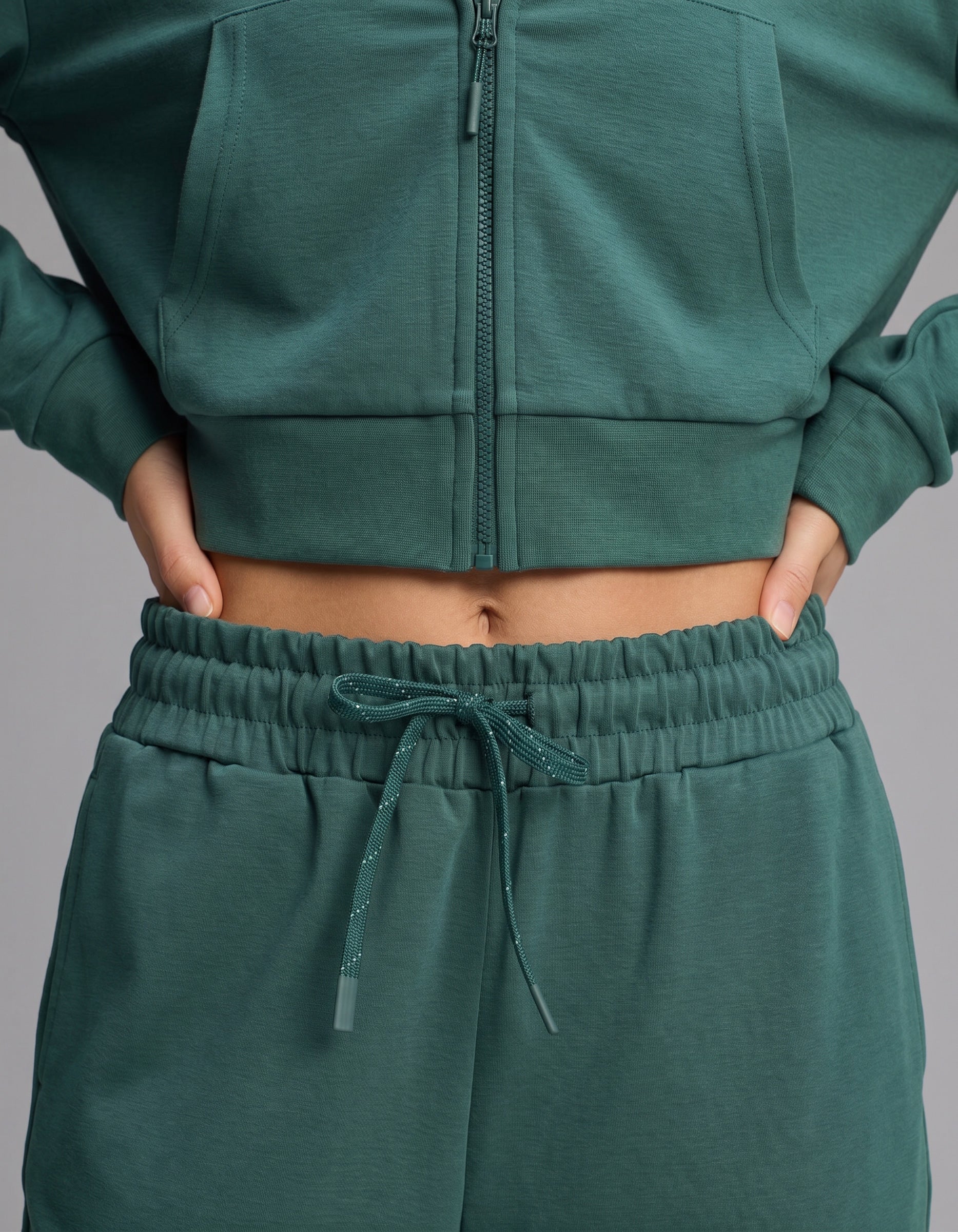 Clad in the Cozy Earth Women's Versa Zip-Down Hoodie & Straight Leg Pant Set in green, a person stands with hands on hips, midriff exposed.