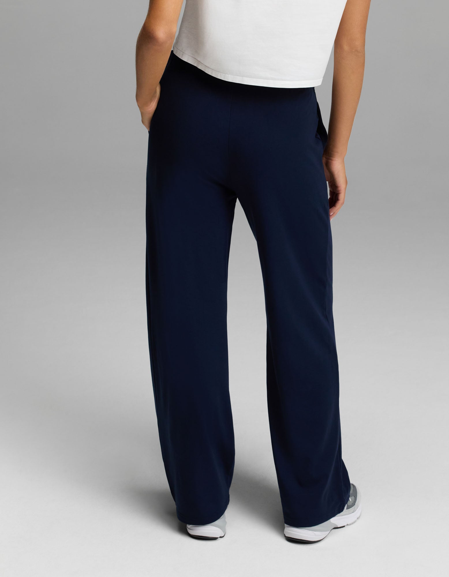 A person stands with their back to the camera against a plain grey background, wearing a white short-sleeve top, white sneakers, and the Cozy Earth Women’s Brushed Bamboo Wide Leg Pull-On Pant in dark blue.