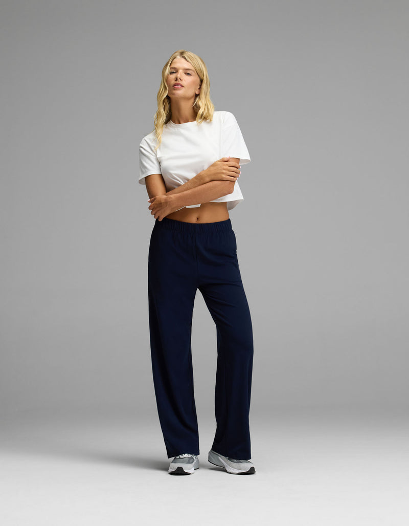 A person stands against a plain background, wearing a white cropped t-shirt, white sneakers, and the Cozy Earth Women’s Brushed Bamboo Wide Leg Pull-On Pant in dark blue. Their long blonde hair falls over their shoulders as they cross their arms and look off to the side.