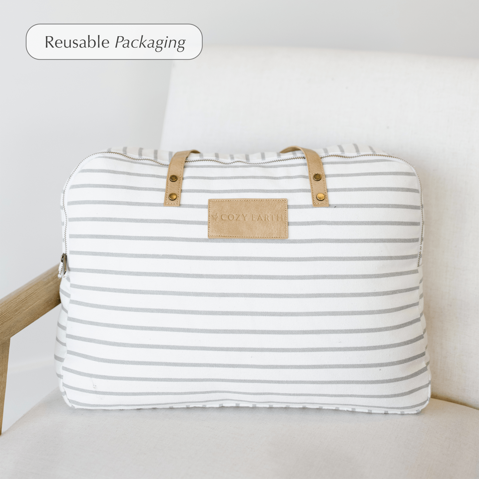 A white, zippered, rectangular bag with light gray horizontal stripes and brown leather handles, labeled "Bamboo Blanket" by Cozy Earth, placed on a white cushioned chair. A label in the top-left corner reads "Reusable Packaging. 