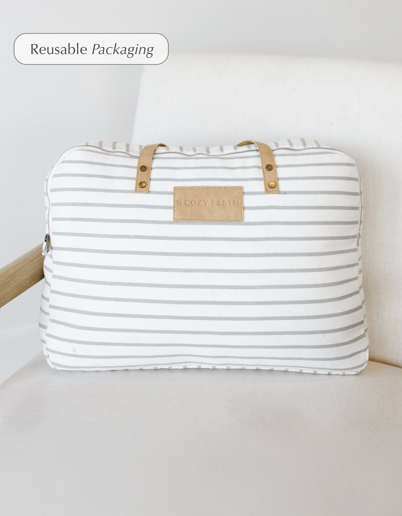 A Cozy Earth Bamboo Blanket in a striped white and gray fabric bag with tan handles and a "COZY EARTH" label rests on a white cushioned chair. A corner label notes the bag is reusable packaging. 