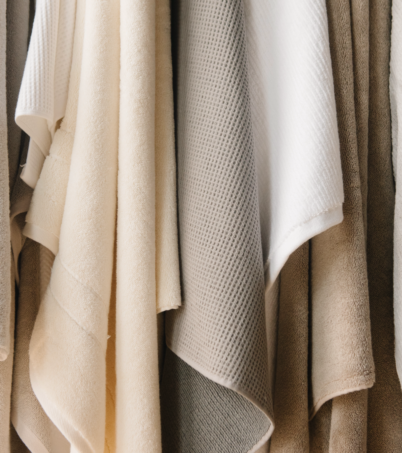 Various textured towels in neutral tones of cream, beige, and gray hang in a row. The materials vary from smooth to waffle-textured, creating a pattern of soft folds and shadows.
