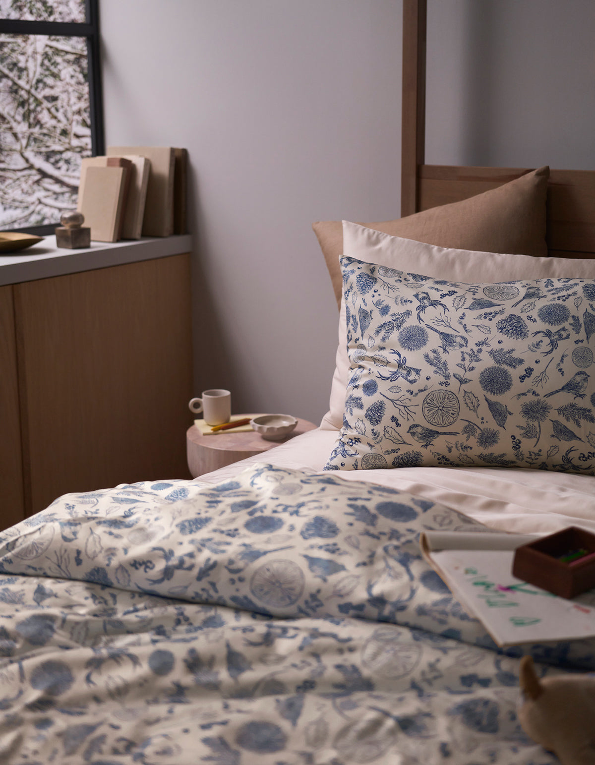A cozy bedroom features a bed topped with the Cozy Earth Bamboo Sheet Set, beige pillows, a small bedside table holding a cup and saucer, books, and a window displaying a snowy scene outside.