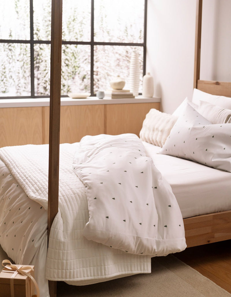 A cozy bed is made with the Cozy Earth Bamboo Sheet Set in white with subtle dark patterns, positioned by a snowy window. Light wood furniture and accents complete the calm, inviting bedroom atmosphere.