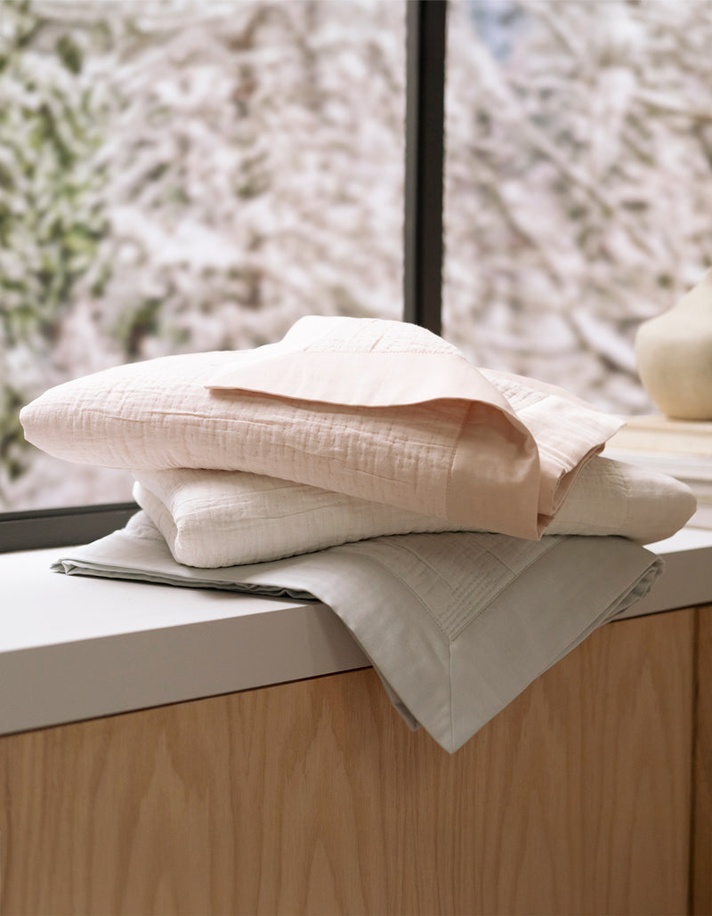 A stack of Cozy Earth's Little Bee Matelasse Baby Blankets in pastel colors rests on a wooden surface near a window, with a softly blurred snowy scene outside.