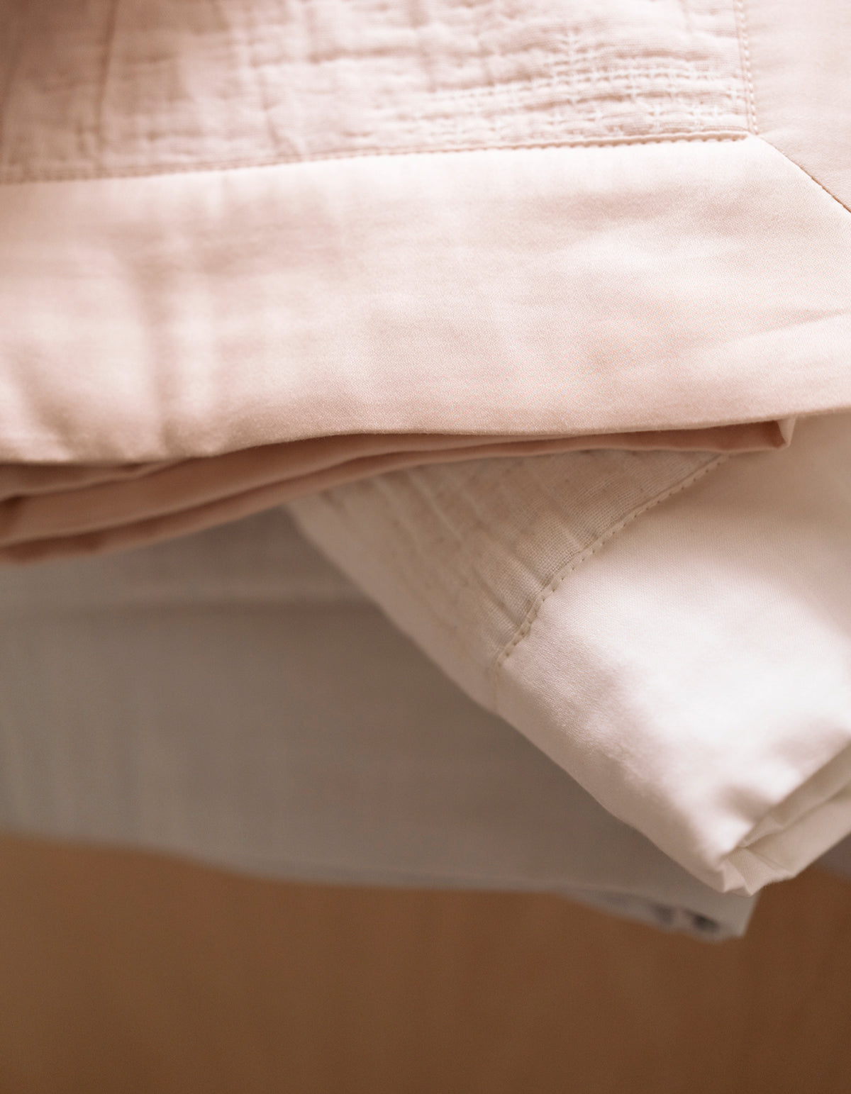A close-up of the Cozy Earth Little Bee Matelasse Baby Blanket shows soft pink and white fabrics neatly folded in a stack, featuring smooth, slightly textured materials.