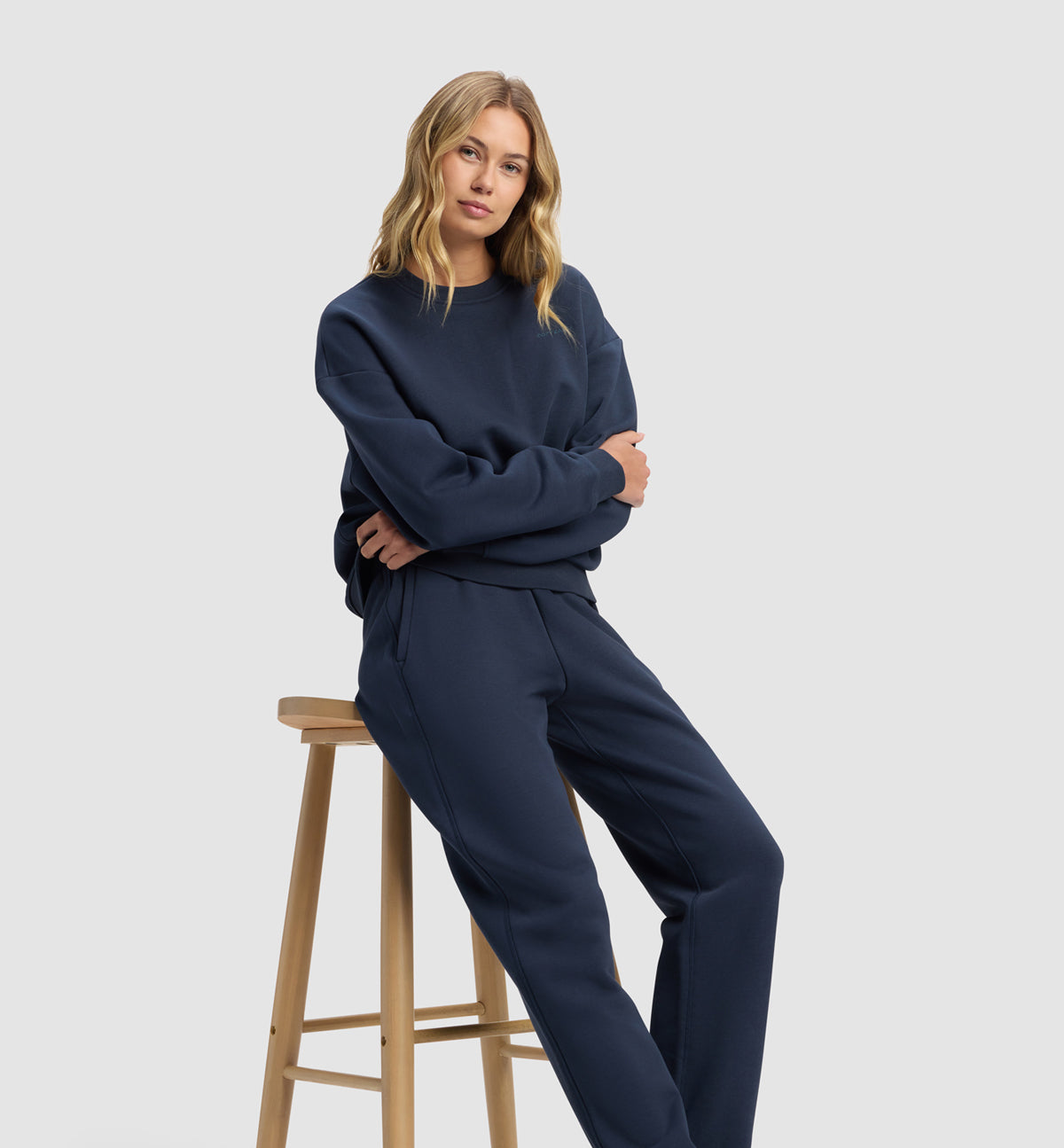 Woman wearing a navy blue Cozy Earth Cityscape 2.0 Crewneck and Jogger Set sitting on a wooden stool against a light gray background