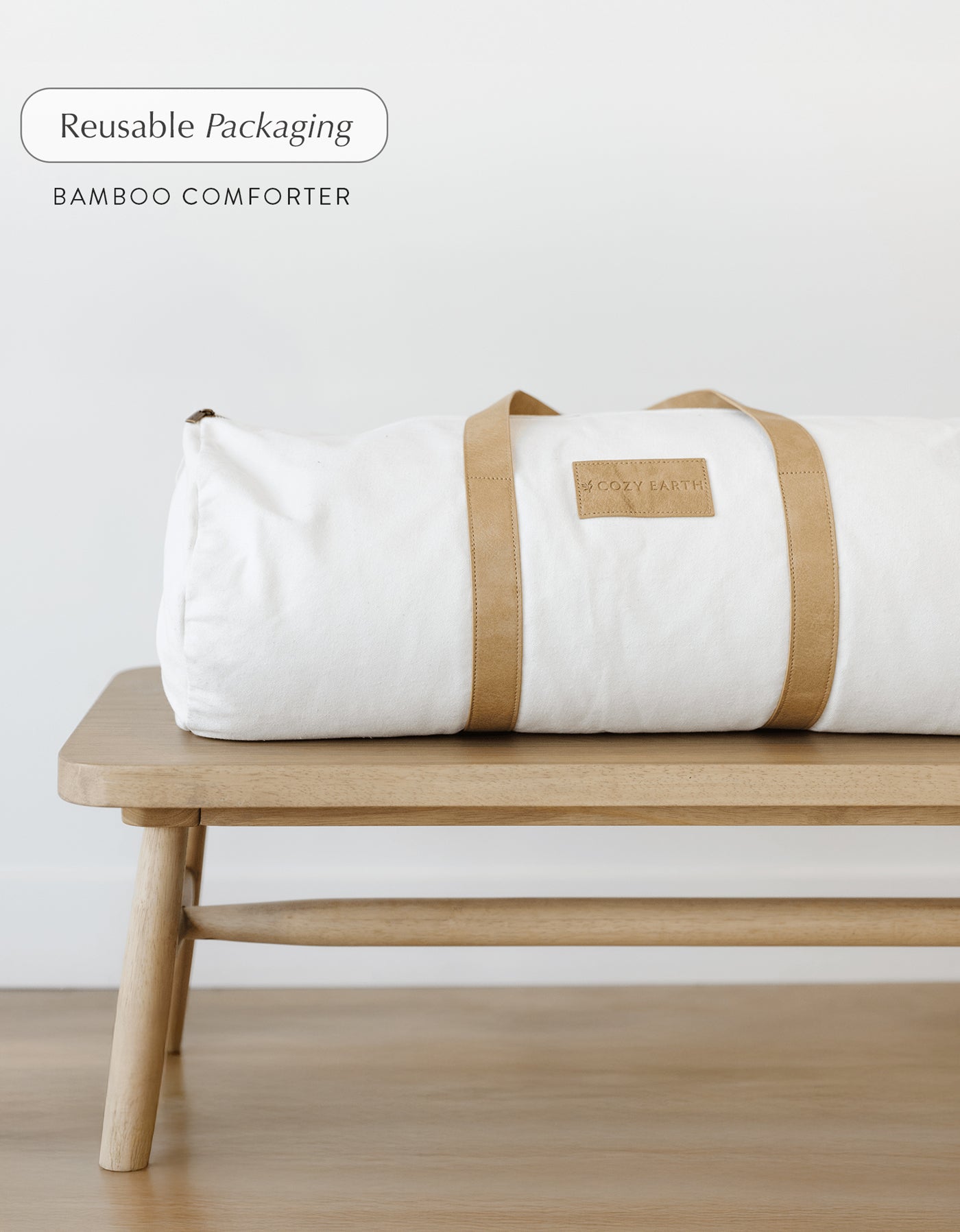 A Cozy Earth Bamboo Viscose Comforter in a reusable duffel bag with tan straps sits on a wooden bench; text reads "Reusable Packaging, Bamboo Viscose Comforter.