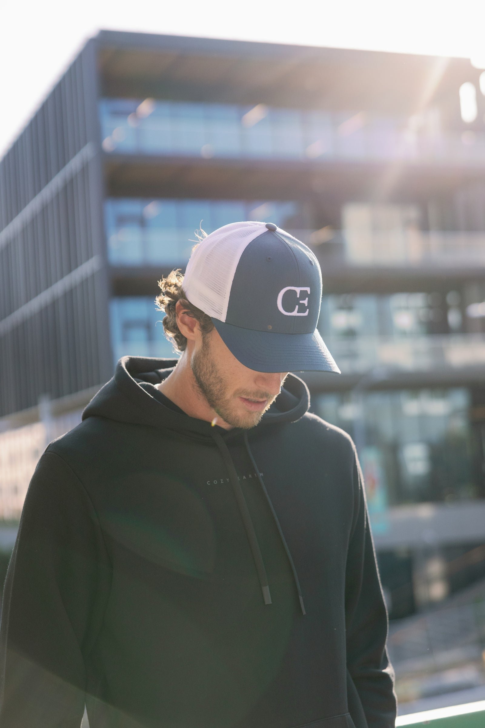A person wearing a blue and white cap and a black hoodie stands outdoors with their head slightly down. A modern glass building is visible in the background, with sunlight casting a soft glow.