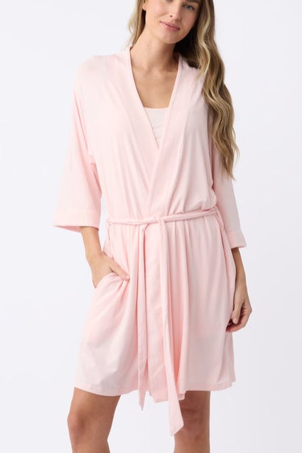 A person wearing a soft pink robe with a matching belt poses against a white background. The robe has three-quarter sleeves and a relaxed fit. The person has long, wavy blonde hair and is smiling gently.