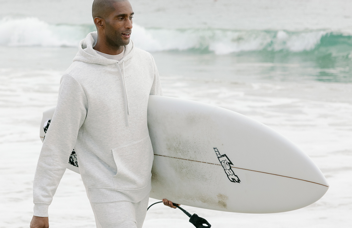 A man carrying a surfboard walks near the ocean wearing Cozy Earth CityScape Hoodie and Sweatpants.