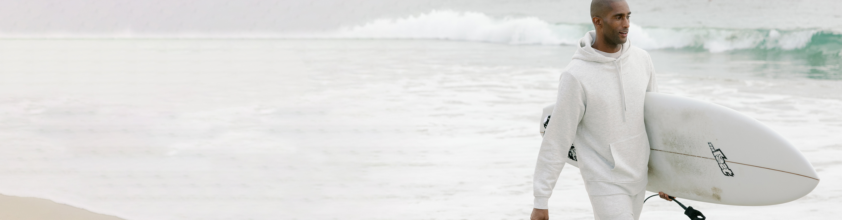 A man carrying a surfboard walks near the ocean wearing Cozy Earth CityScape Hoodie and Sweatpants.
