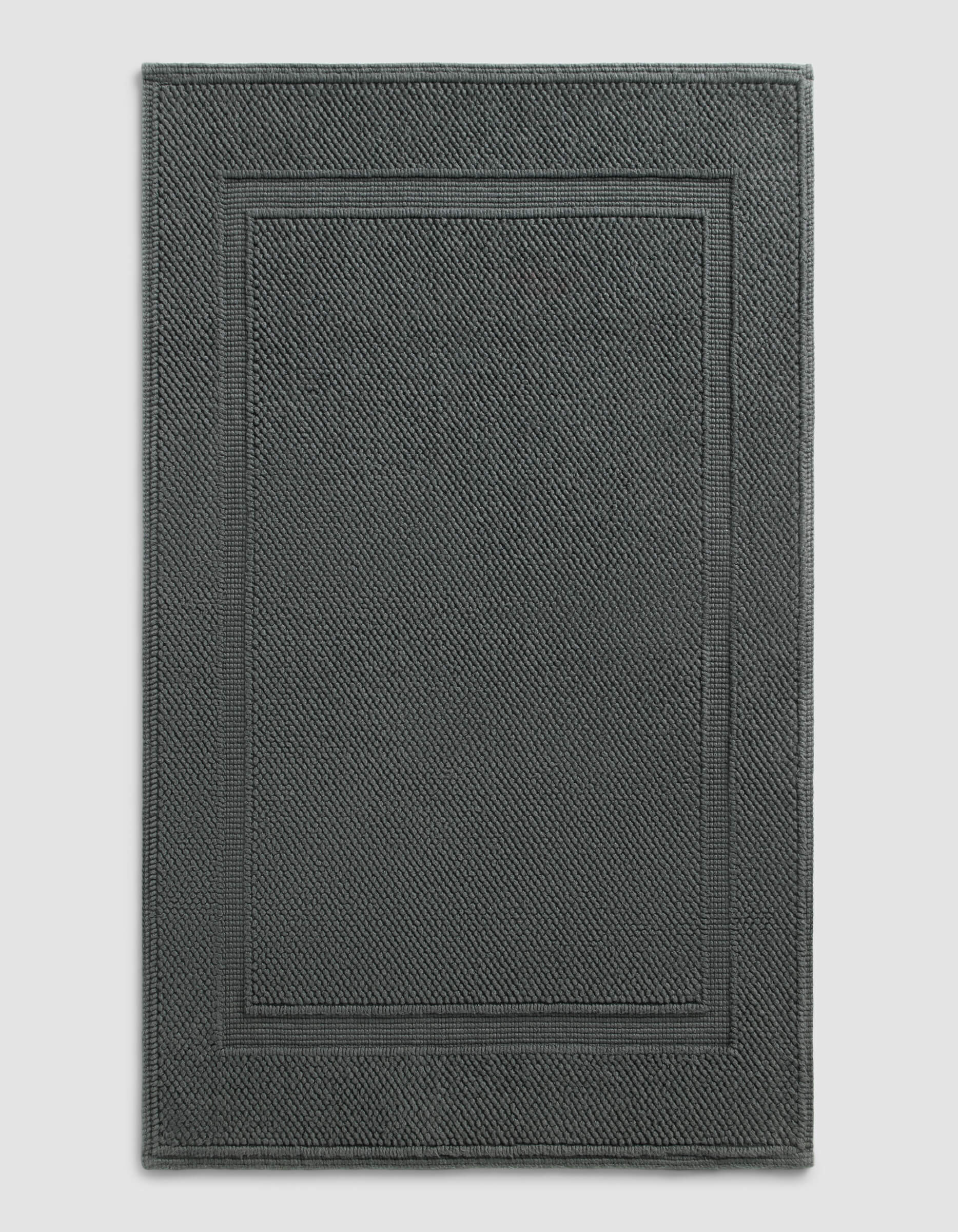 The Cozy Earth Classic Bath Mat features a dark gray, rectangular design with a textured surface and double border, shown against a plain light background.|Color:Graphite