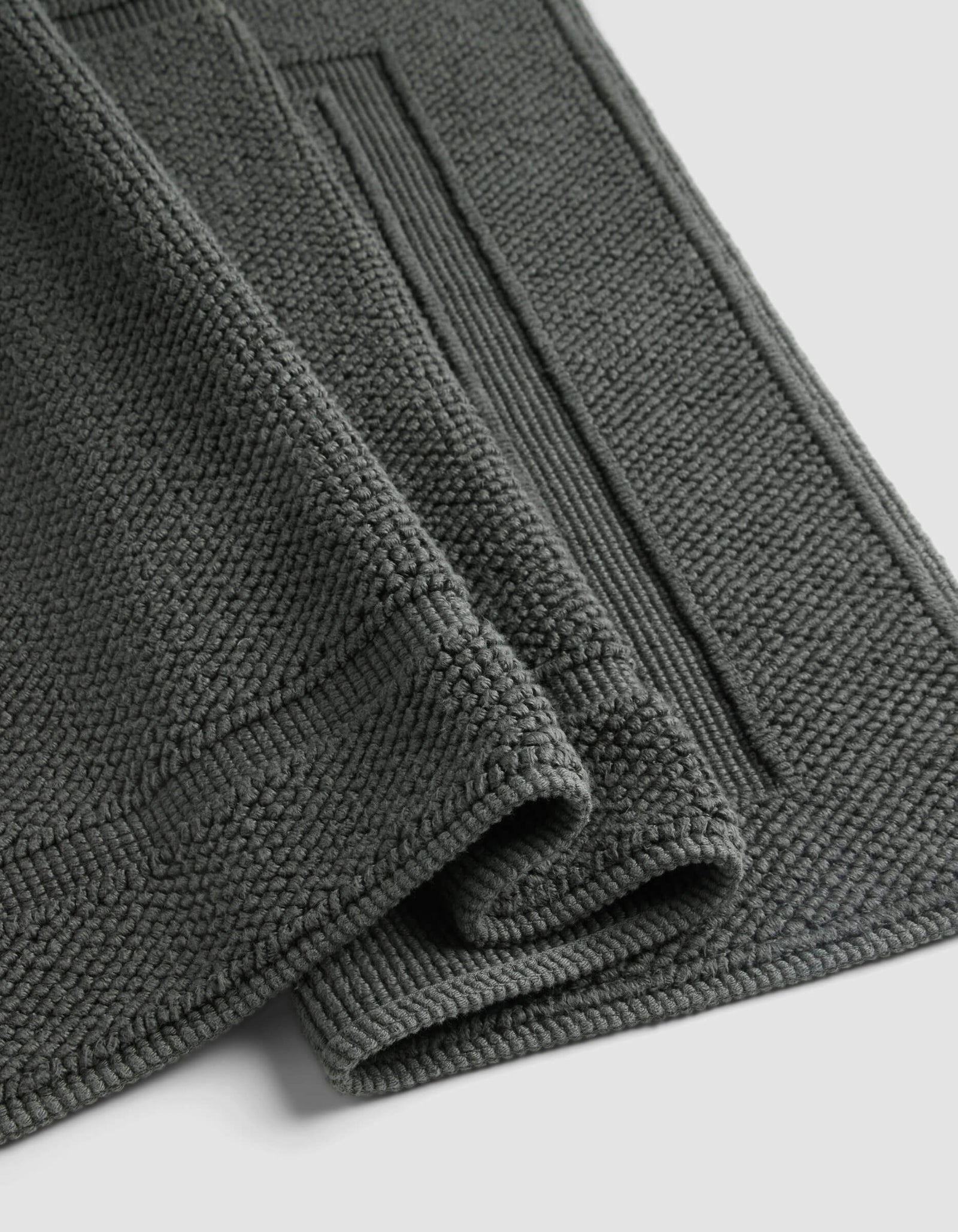 The Cozy Earth Classic Bath Mat, shown folded against a light background, features a dark grey textured finish with a thick, soft pile and a rectangular border pattern.