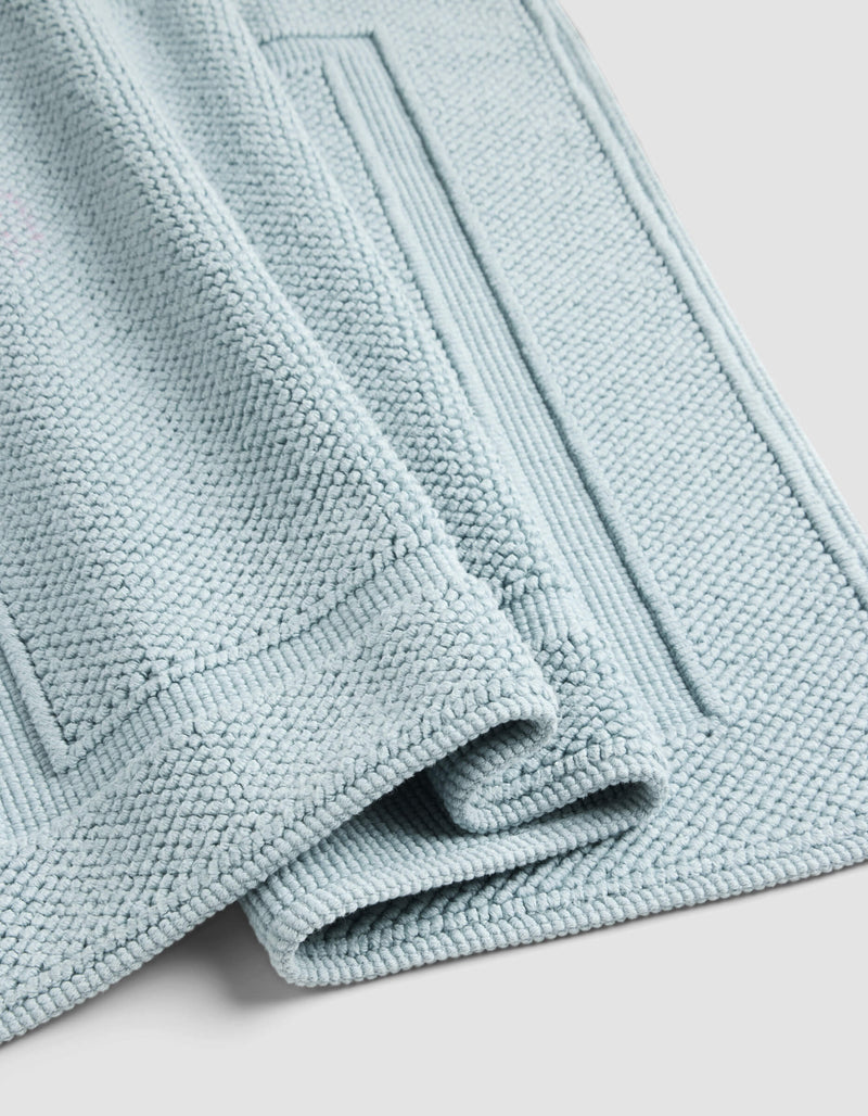 A close-up of the Cozy Earth Classic Bath Mat in light blue, neatly folded to display its textured surface and rectangular border patterns, set against a light gray background.