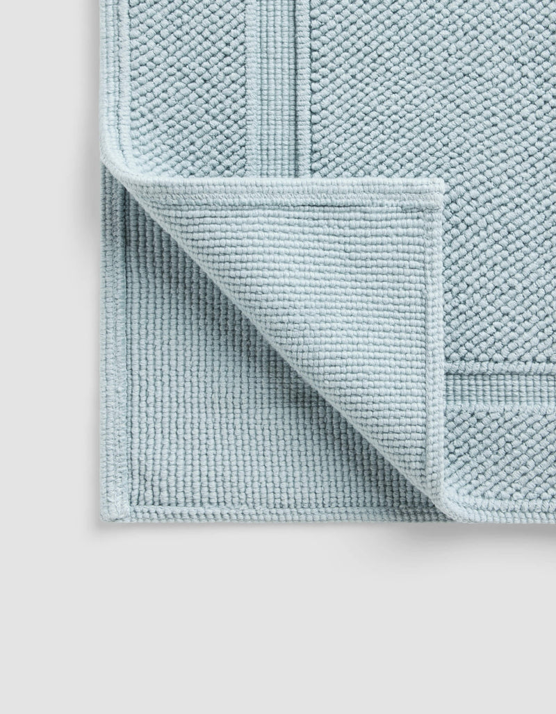 The Cozy Earth Classic Bath Mat, in light blue with a textured surface and border pattern, is shown with one corner flipped to display the underside, resting on a plain, light-colored background.