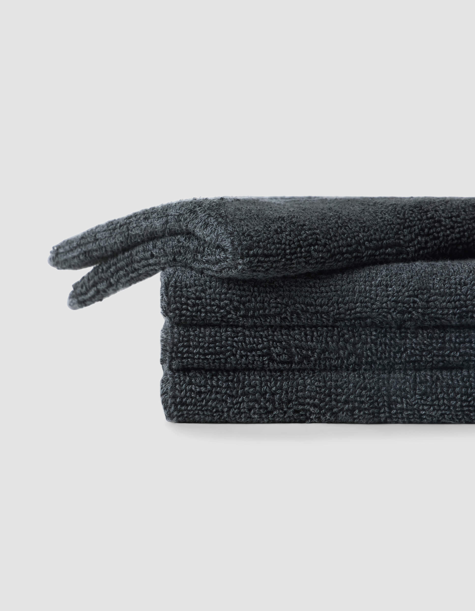 A stack of four neatly folded Complete Cleansing Cloths by Cozy Earth, in dark gray, sits on a light gray background. The towels have a soft, plush texture.