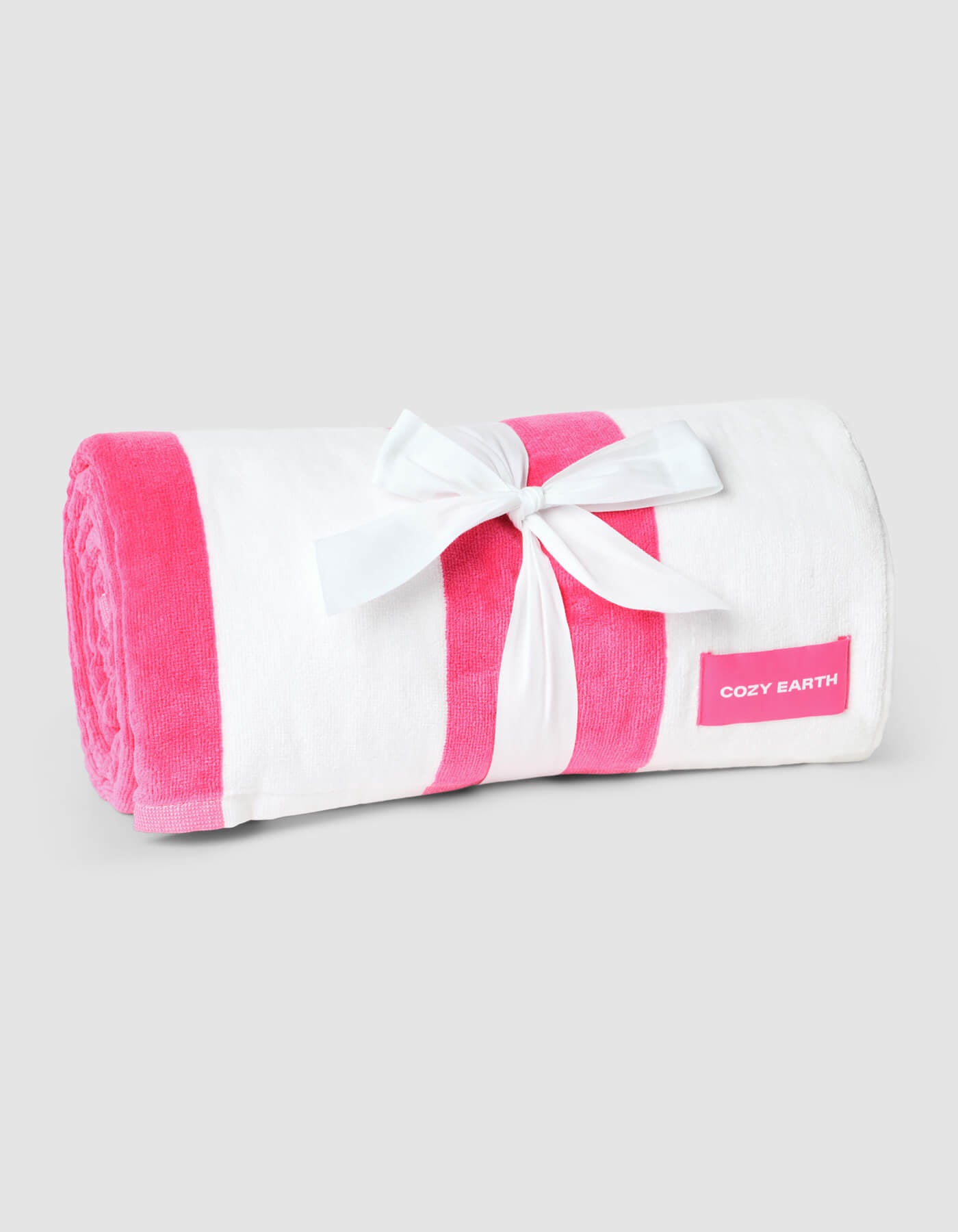 The Seaside Resort Towel by Cozy Earth is a rolled-up white towel with pink stripes, tied with a white ribbon bow and a pink “Cozy Earth” label on the side, displayed against a plain light gray background.