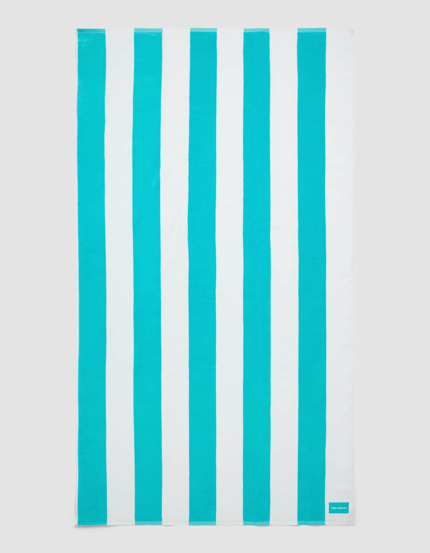 The Cozy Earth Seaside Resort Towel features alternating vertical turquoise and white stripes with a small turquoise label stitched on one corner, set against a light gray background.