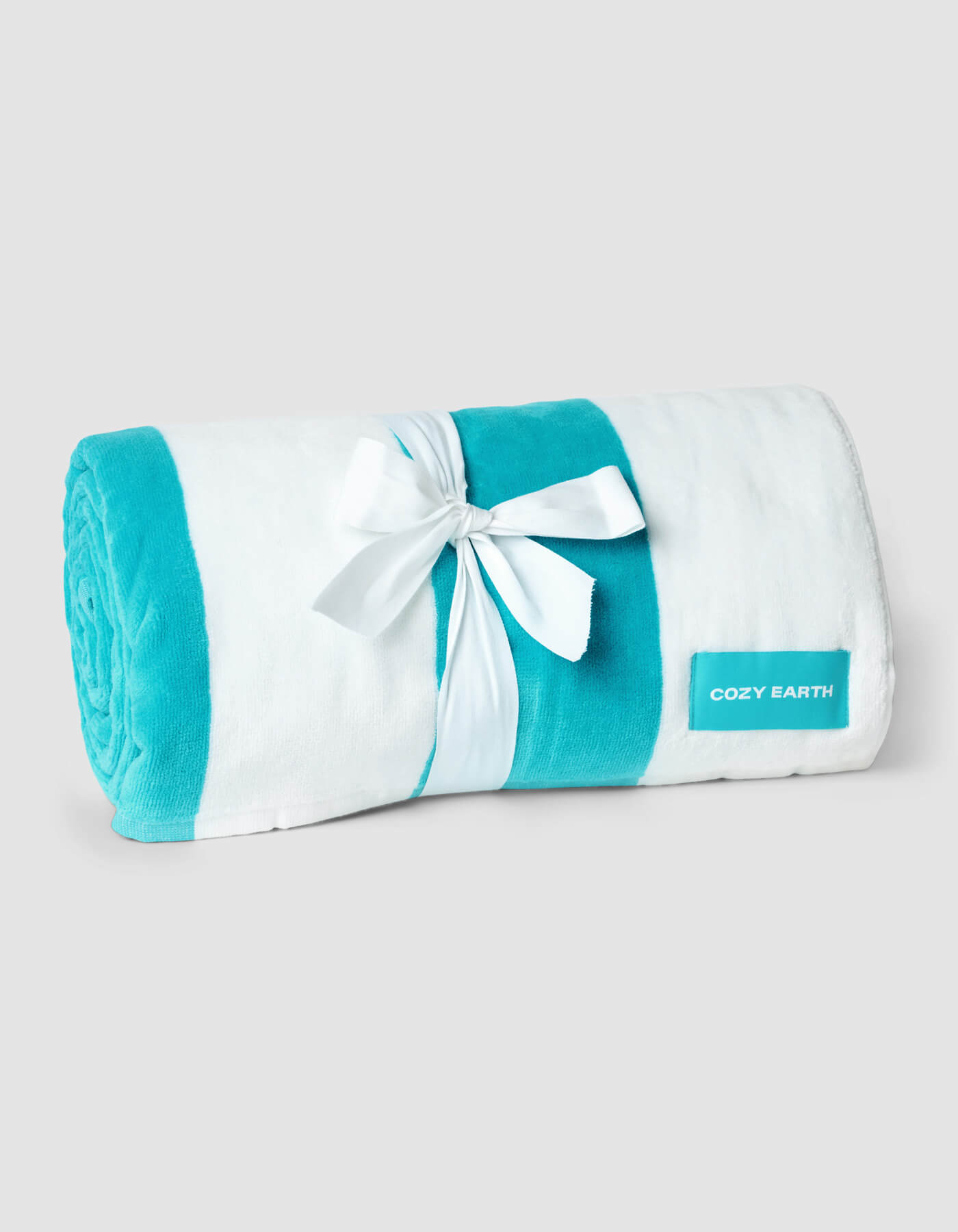 A rolled-up white and turquoise Seaside Resort Towel by Cozy Earth is tied with a white ribbon bow. A turquoise label with "Cozy Earth" is attached to the towel. The background is a plain light gray.