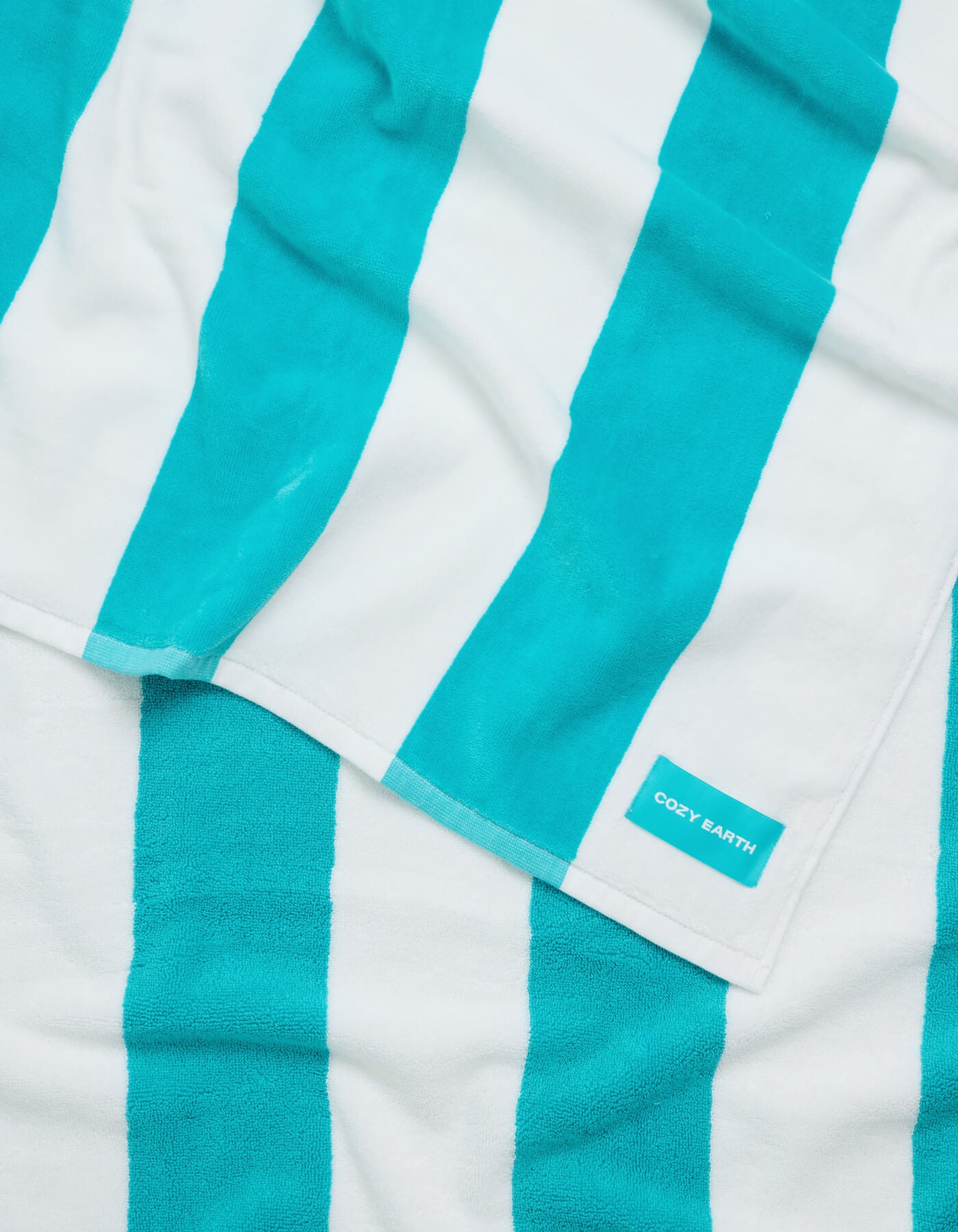 A neatly folded Seaside Resort Towel by Cozy Earth, featuring turquoise and white stripes with a visible "COZY EARTH" label, rests atop another matching towel.|Color:Aqua Stripe