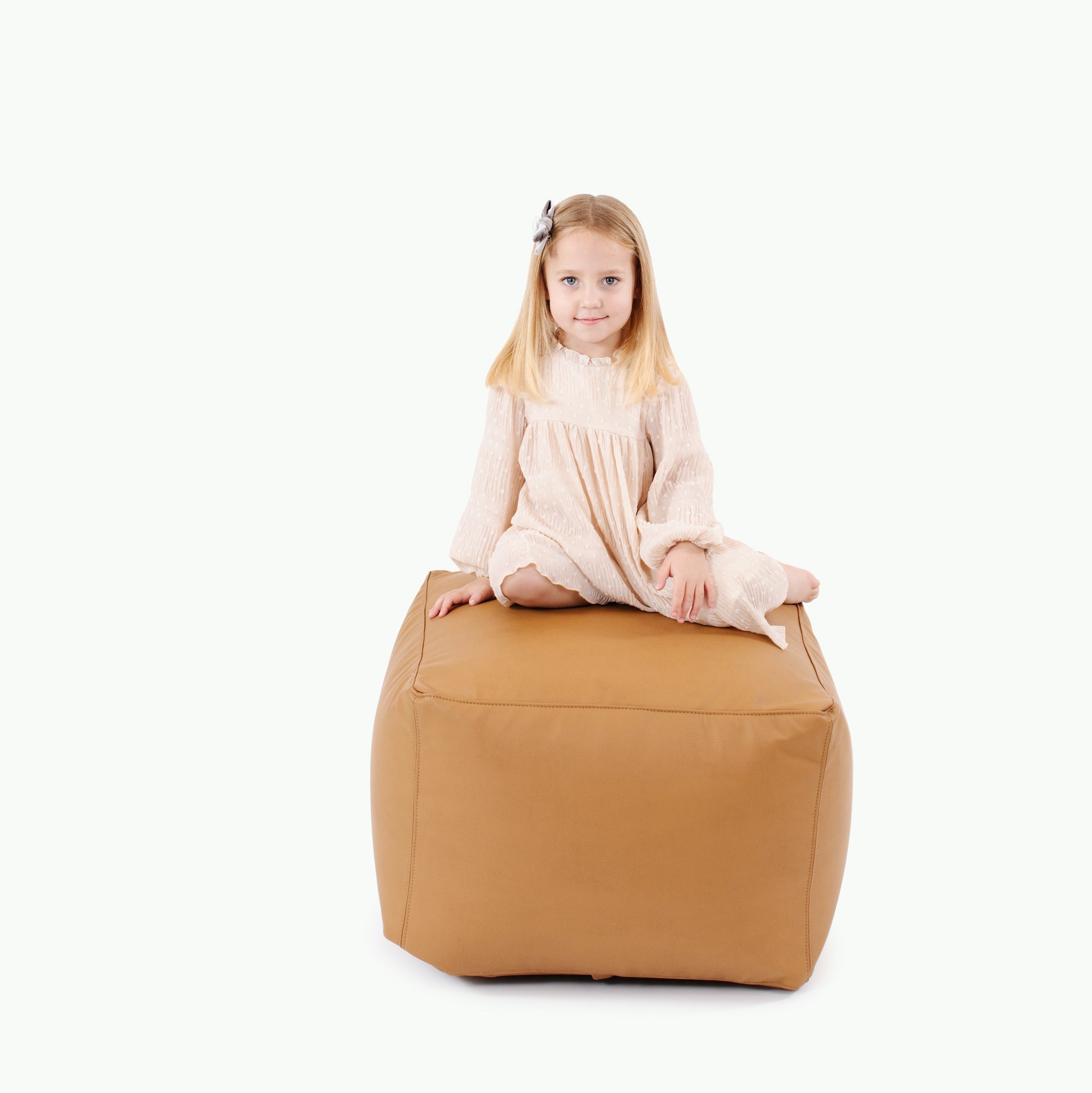 A young girl with blonde hair, in a light dress and hair clip, sits cross-legged on the Gathre Wholesale Pouf in Camel against a plain white background.