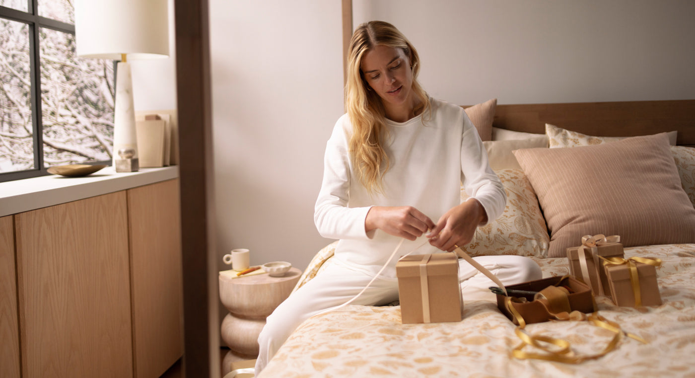Woman in a bedroom setting preparing a gift with decorative ribbons.