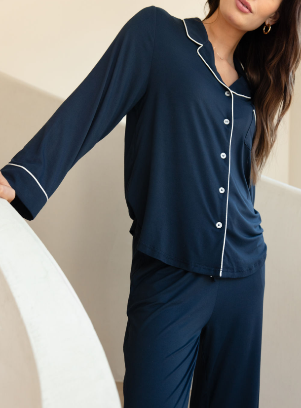 A woman with long brown hair, partially visible, leans on a white curved surface. She is wearing Cozy Earth's Women's Stretch-Knit Long Sleeve Bamboo Pajama Set in dark blue, featuring white piping on the collar, cuffs, and button lining. The background is neutral and minimalist. 