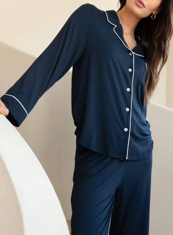 A woman with long brown hair, partially visible, leans on a white curved surface. She is wearing Cozy Earth's Women's Stretch-Knit Long Sleeve Bamboo Pajama Set in dark blue, featuring white piping on the collar, cuffs, and button lining. The background is neutral and minimalist. |Color:Navy