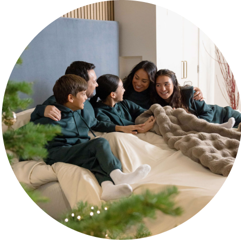 A family of two parents and three children wearing matching green Cozy Earth CityScape Hoodie and Sweatpant Sets cuddle in a bed with a Cozy Earth Cuddle Blanket and Christmas tree in the foreground.