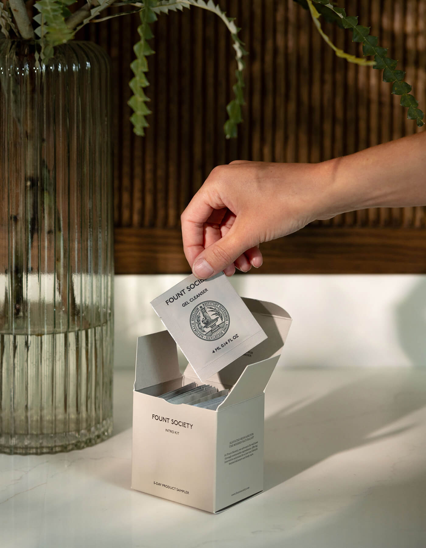 A hand takes the Fount Society Intro Kit by Cozy Earth from a white box on a table beside a tall, ribbed glass vase with leafy green stems.