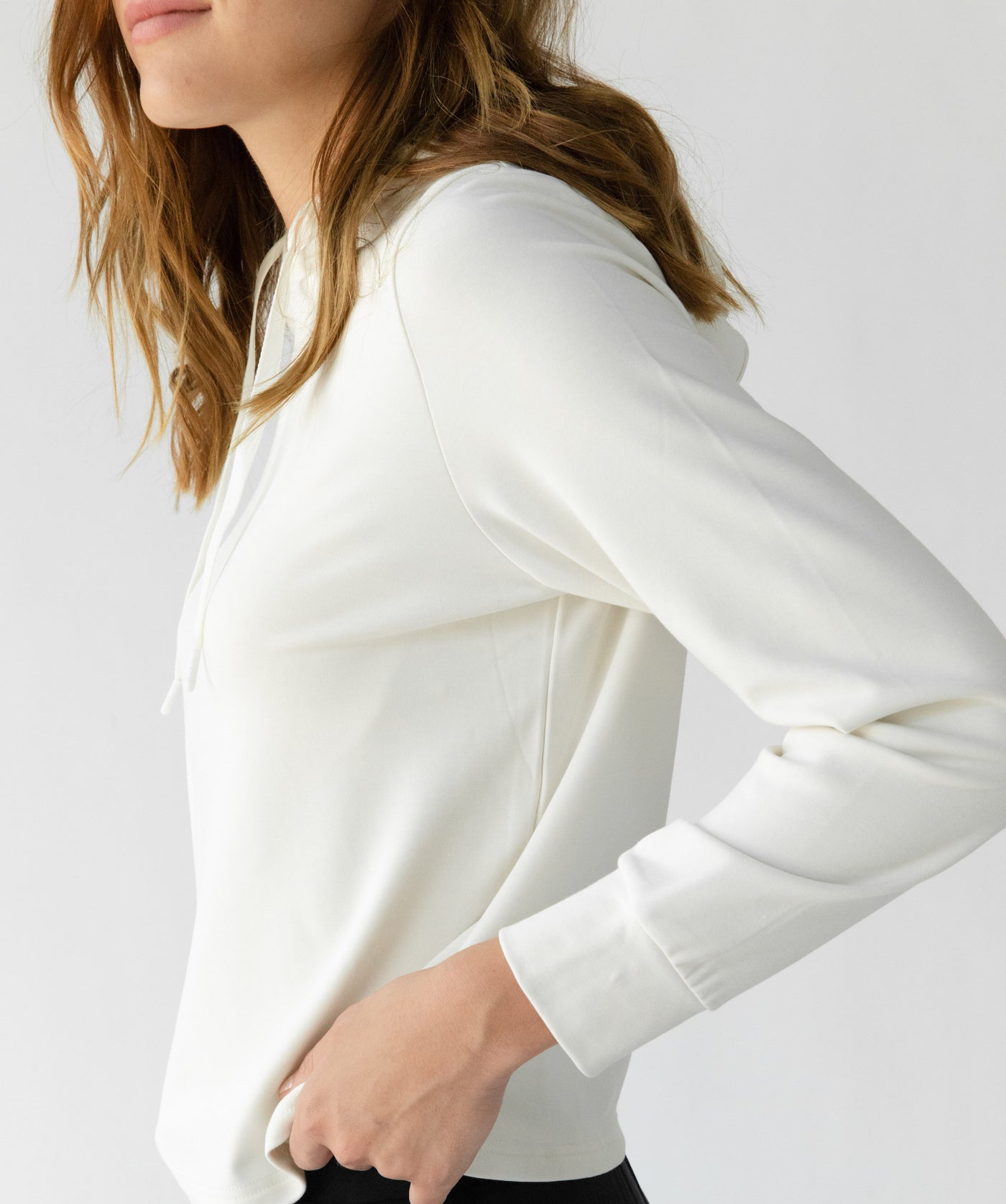 A person with wavy hair is wearing Cozy Earth's Women's Ultra-Soft Bamboo Wide Leg Pull On Pant & Hoodie Set in white, featuring a long-sleeve hoodie and black pants. The image captures a side profile of their torso and part of their face against a neutral background. 