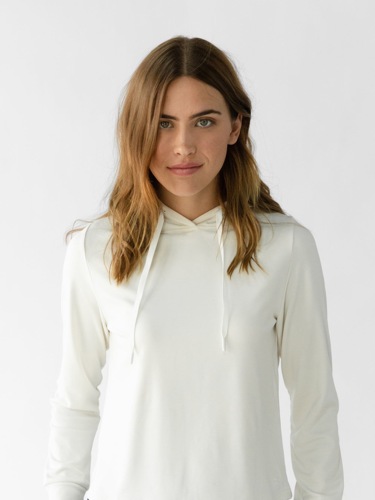 A person with long brown hair is wearing a white hoodie from the Cozy Earth Women's Ultra-Soft Bamboo Wide Leg Pull On Pant & Hoodie Set. They are standing against a plain white background, looking directly at the camera with a neutral expression. 