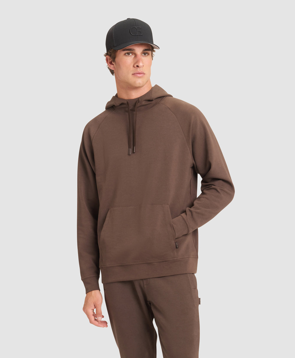 A man wearing a black baseball cap and brown Cozy Earth StretchTech Hoodie and Jogger set stands in front of a grey background.