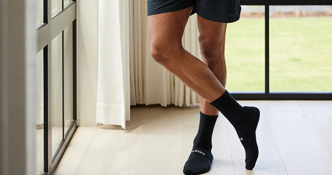 A person wearing black socks and dark shorts stands indoors on a wooden floor in front of a large window with curtains. One leg is slightly crossed over the other, and outside, green grass is visible.